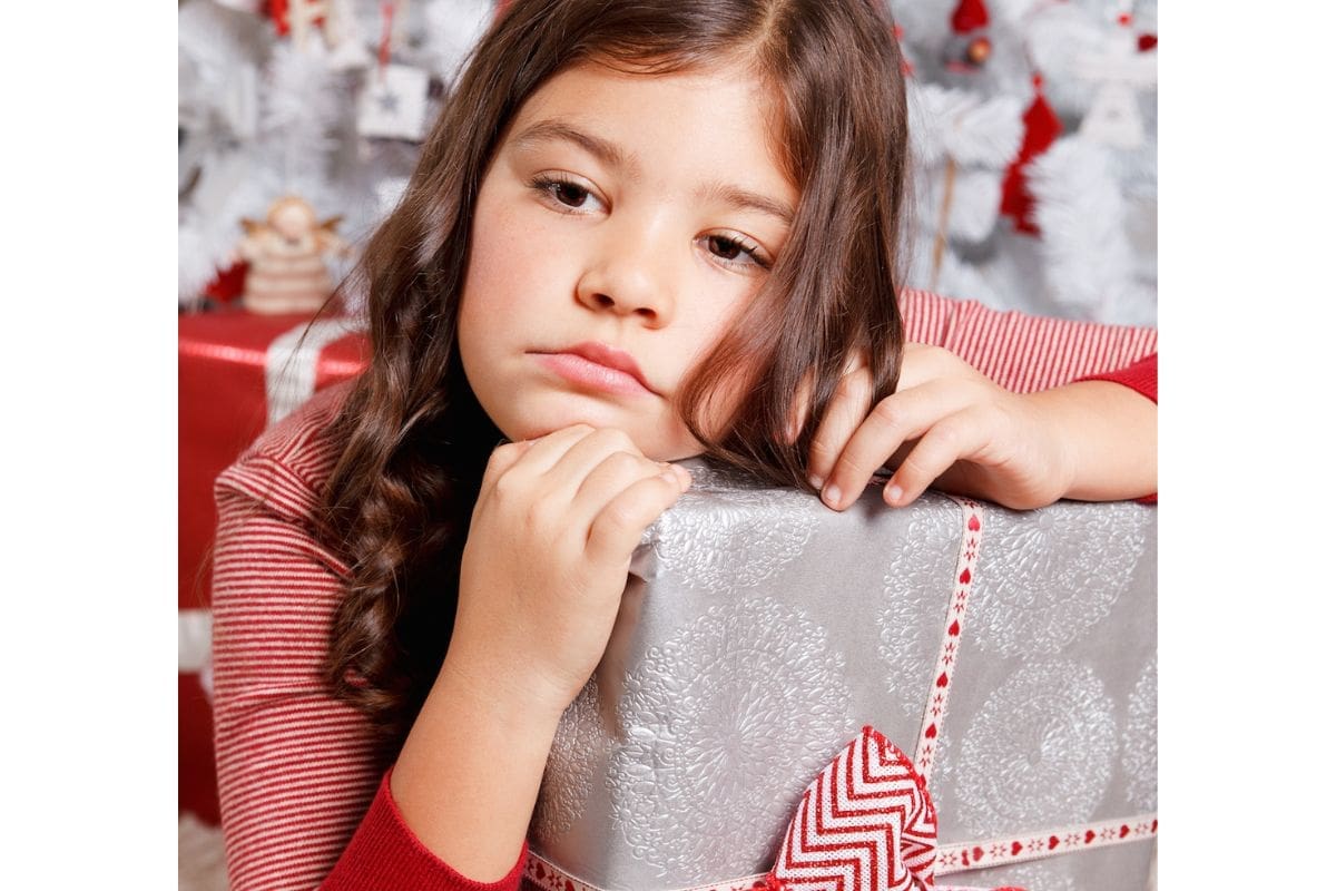 A sad little girl holding a present in front of a white Christmas tree.