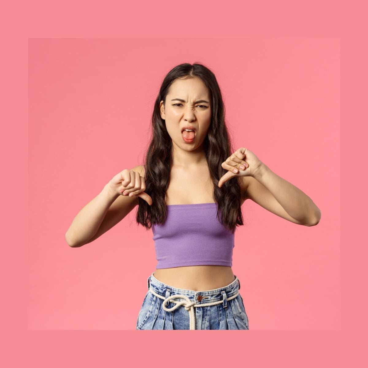 Portrait of disappointed young attractive woman with her thumbs-down and tongue out, grimacing from aversion and dislike, disappointed, pink background.