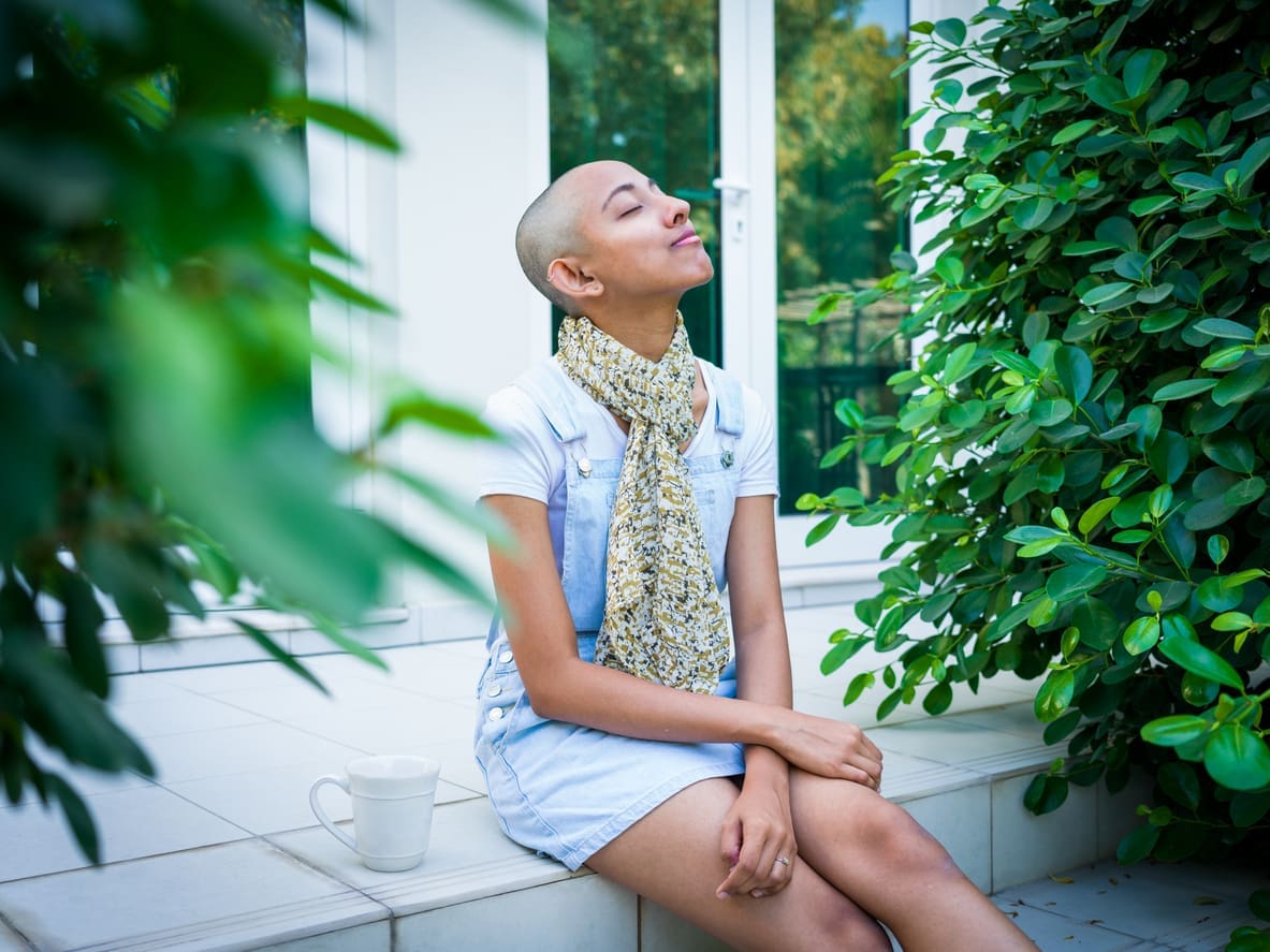 Woman with shaved head sitting outside with cup of coffee looking up to sky in contentment, being resilient