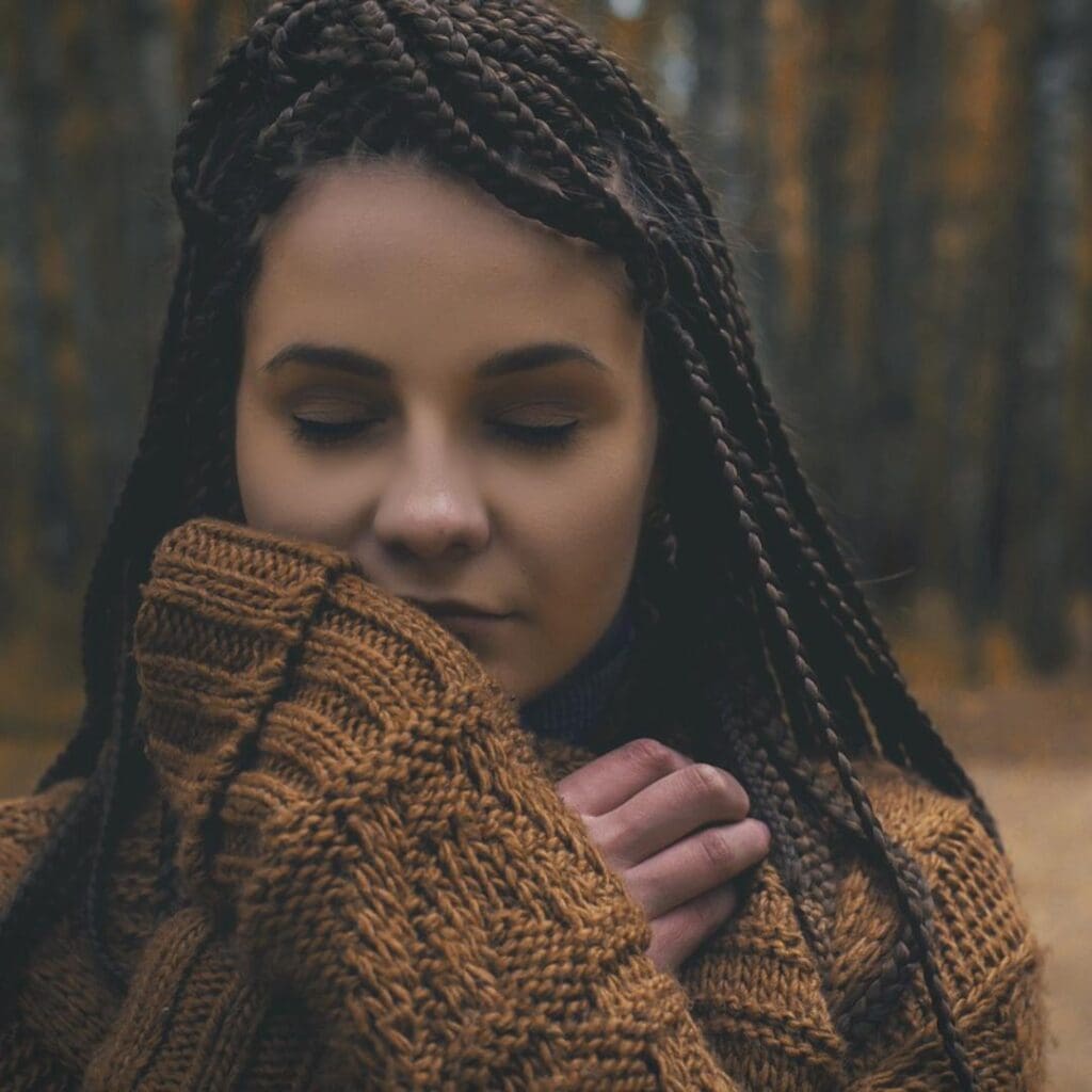 Black/Indigenous woman with long dark braids and cozy sweater in woods eyes closed looking peaceful and happy practicing mindfulness 