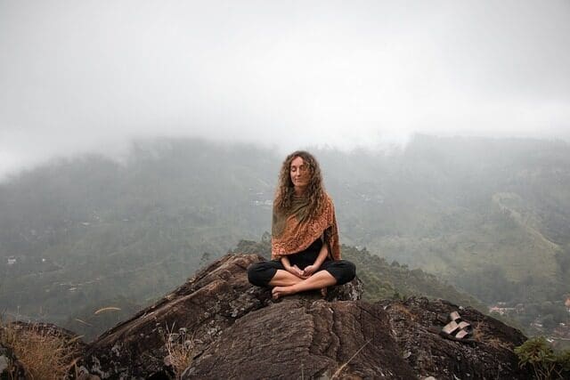 Woman sitting on a rocky hill in front of a cloud-shrouded mountain practicing mindfulness 