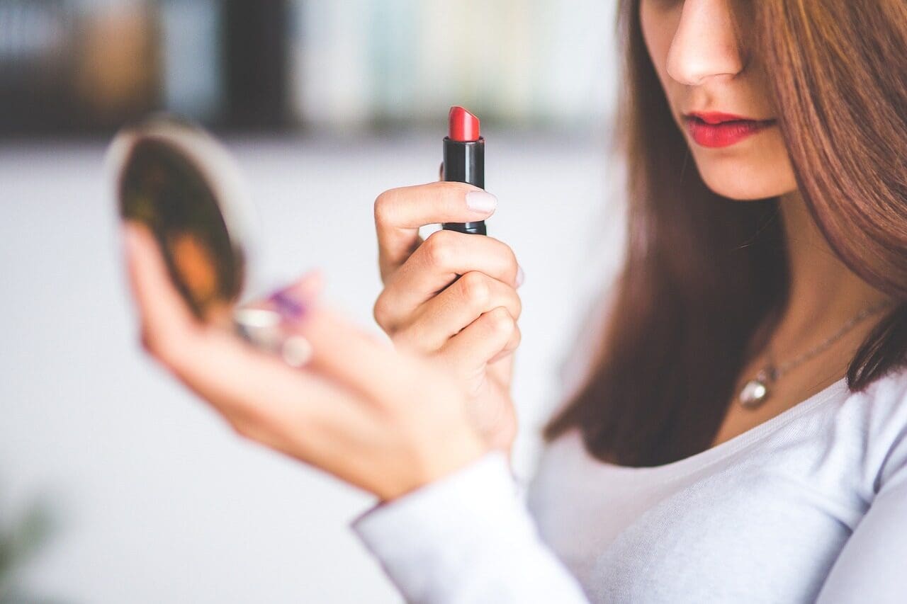 Woman holding a tub of bright red lipstick looking in a compact mirror at her red lips. 