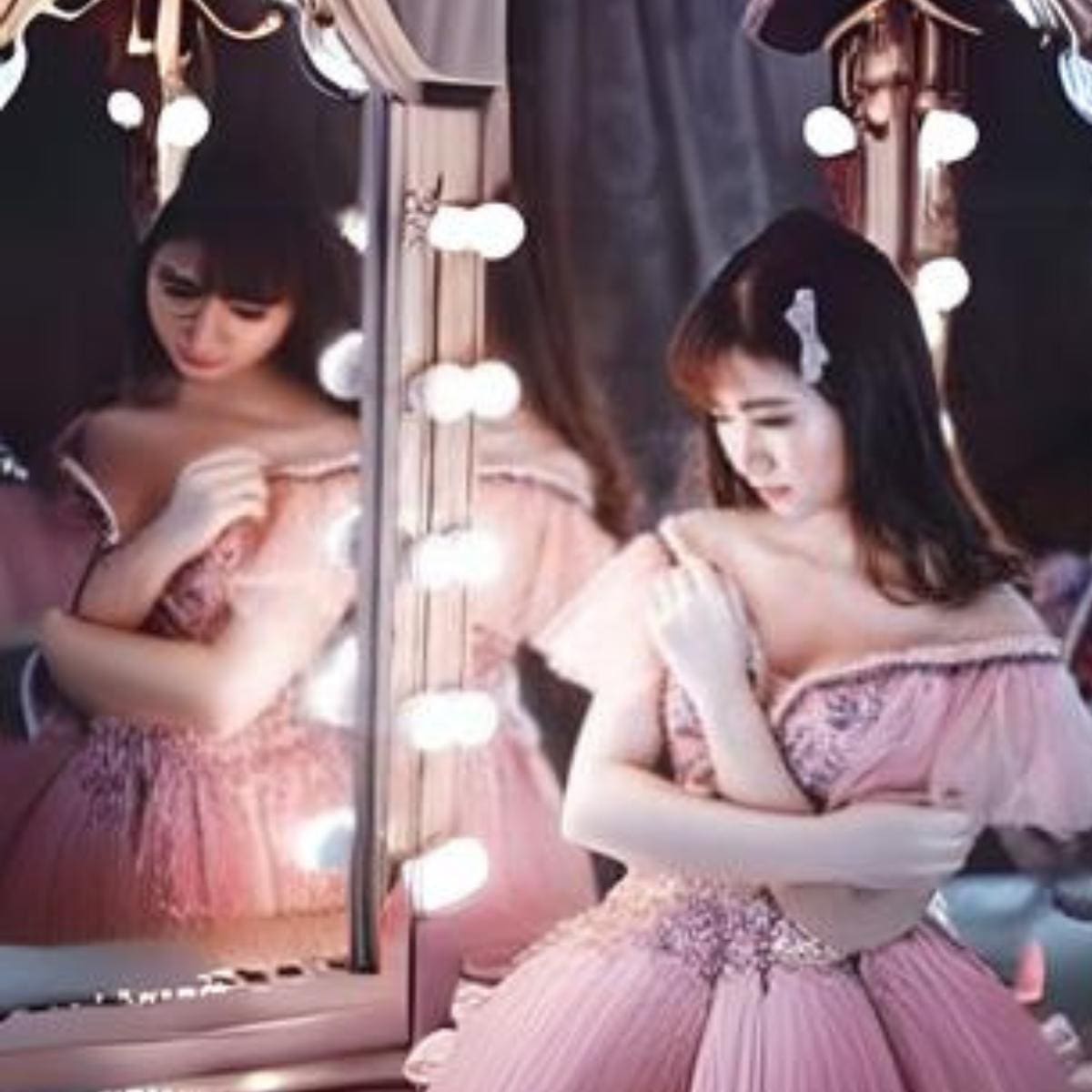 Asian woman dressed in pink dress looking in mirror with bright lights all around.