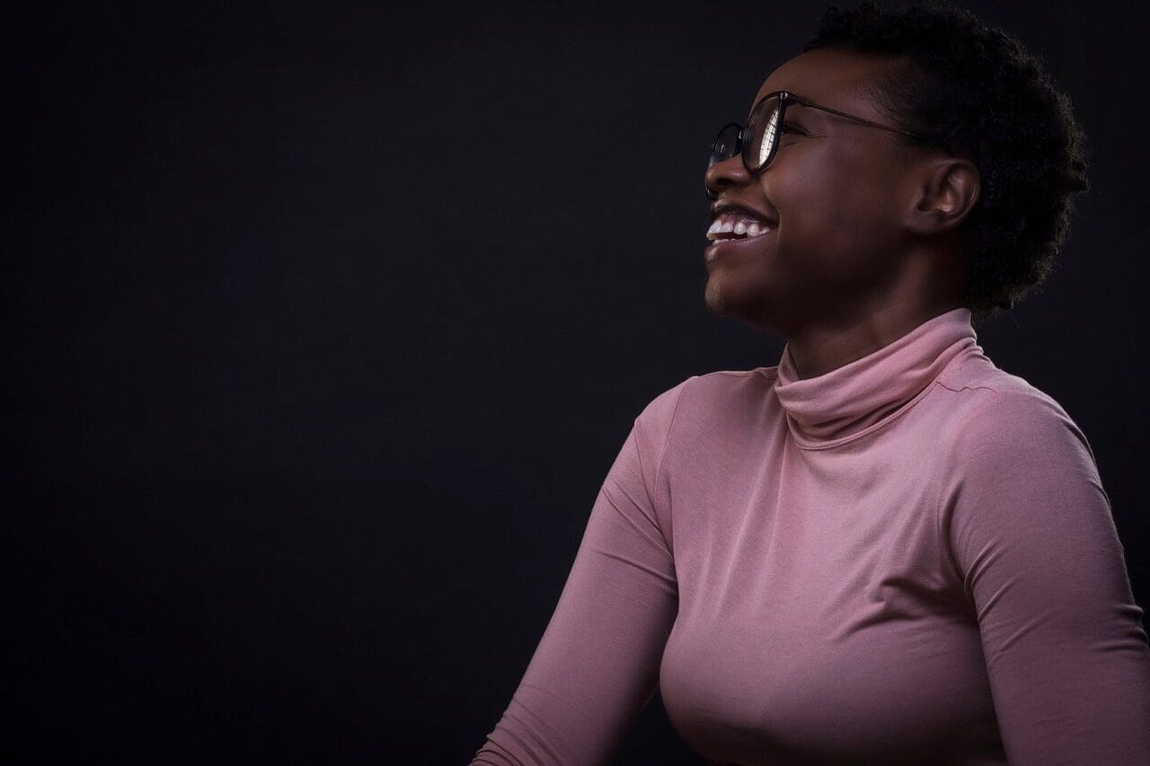 Profile of an African American woman wearing a pink turtleneck and dark-rimmed glasses smiling/laughing 