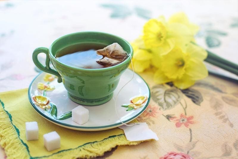 Green teacup on a ceramic saucer with steeping herbal tea inside, a few sugar cubes and a bouquet of daffodils sit next to the cup. 