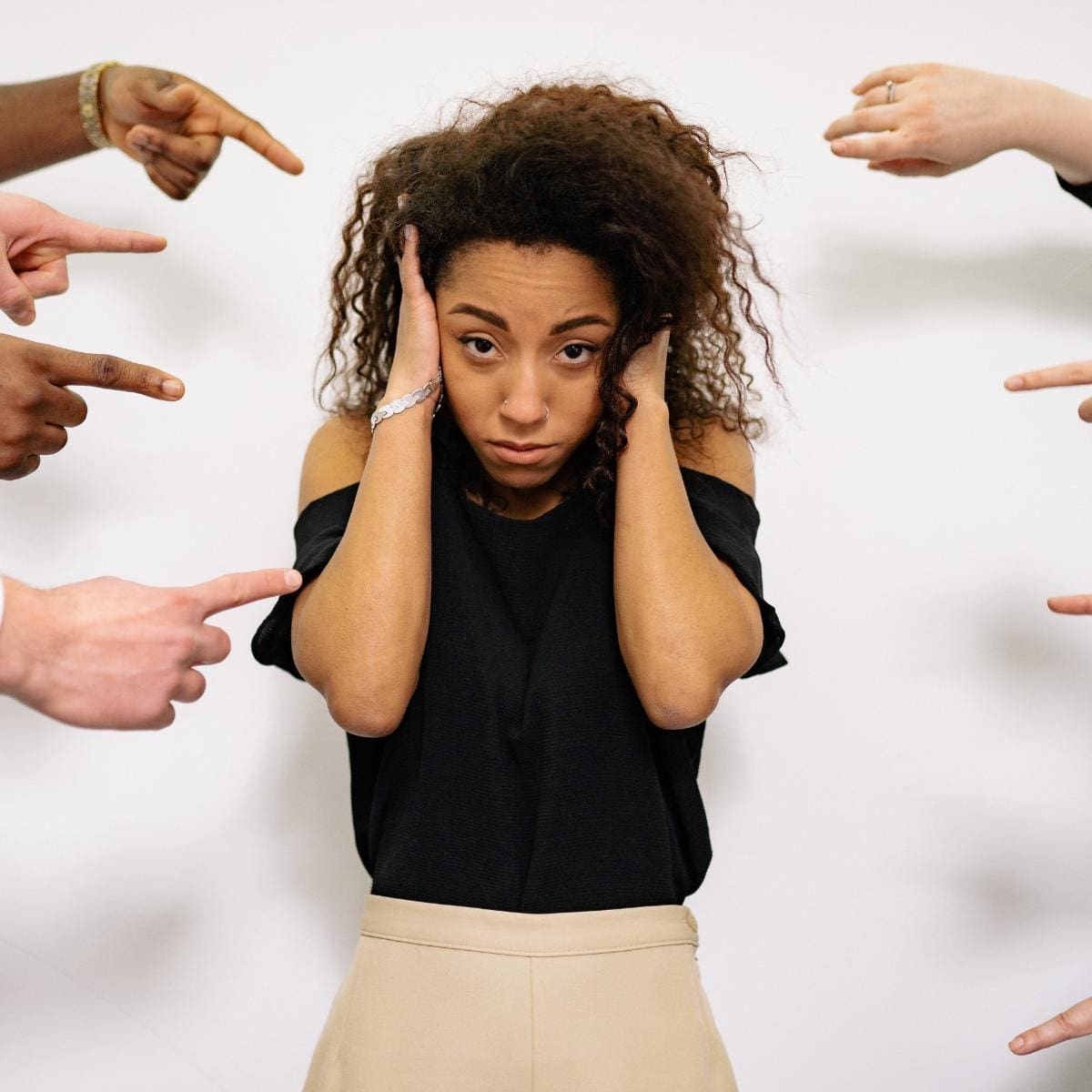 A woman of color being pointed at by various fingers and covering her ears.