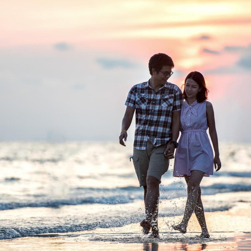 Asian-American couple walking hand-in-hand along the beach at sunset, looking happy and romantic.