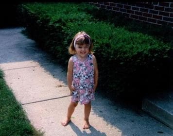 Kristin Oesterle as a very young girl, standing in front of shrubbery of a home in a floral jumper. 