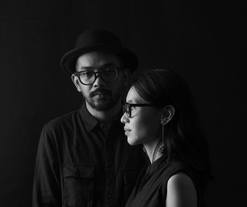 Black and white image of man and woman, both wearing glasses, standing perpendicular to one another, looking somber