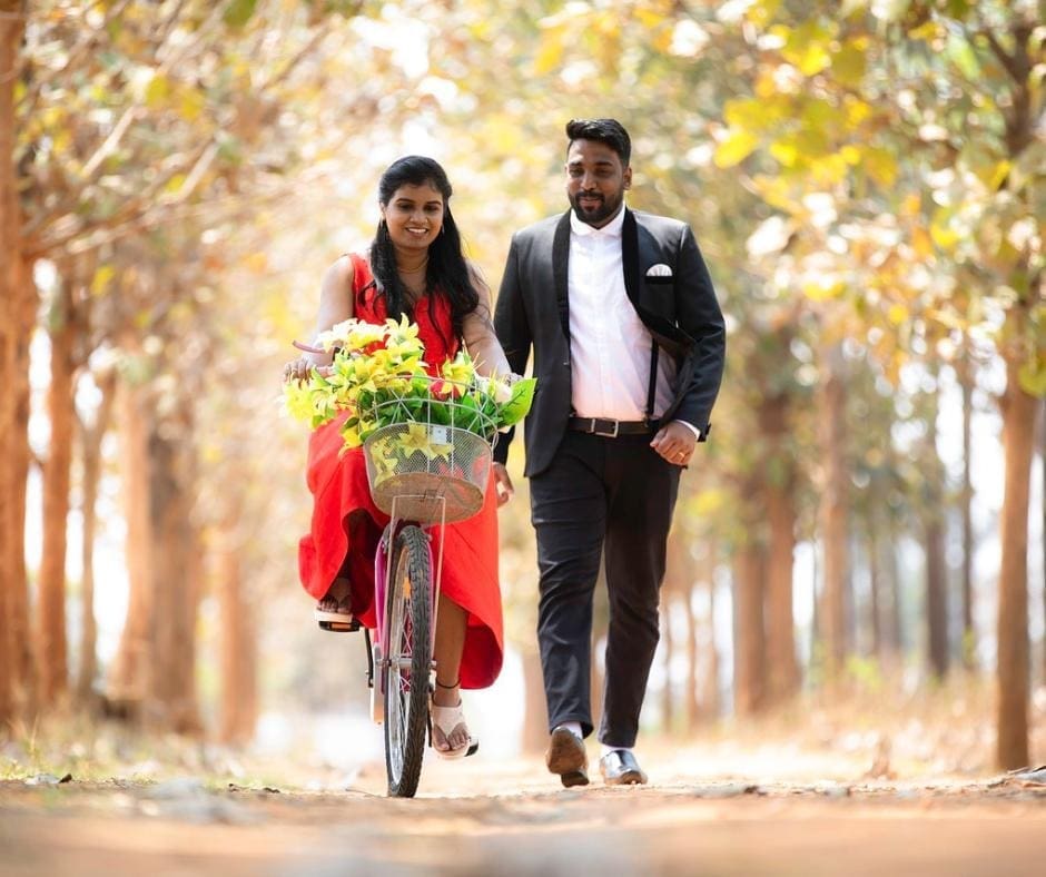 Indian couple together in a bright, tree-lined path. The woman is wearing a red dress and riding a bike with a basket full of yellow flowers, while the man walks beside her in a black and white suit. 