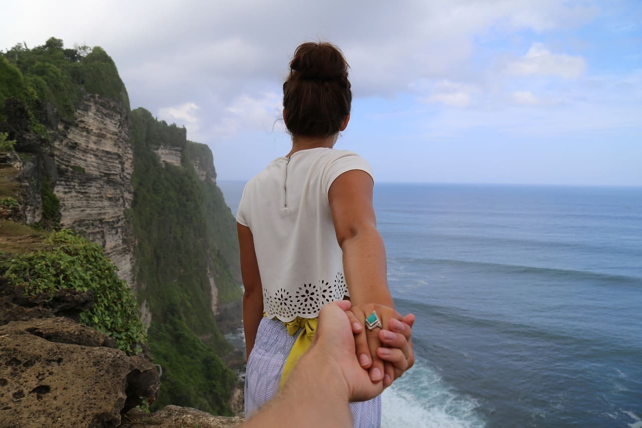 Woman standing near a cliff looking at the ocean, holding the hand of someone behind her that she is not facing/looking at.