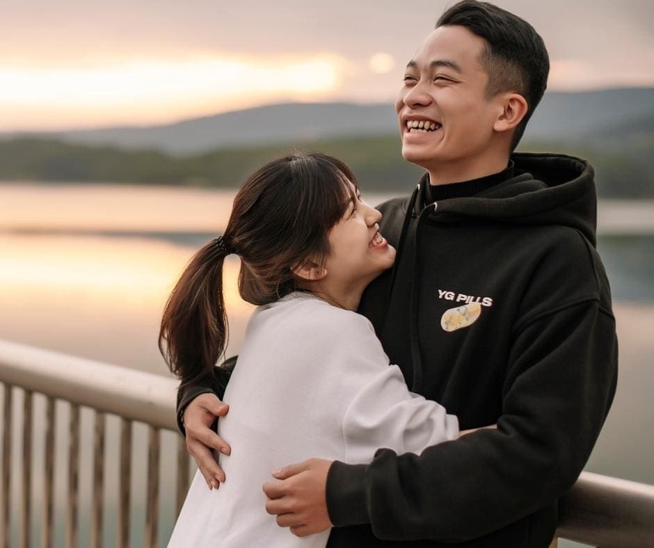 Asian couple hugging and laughing on a bridge over a serene lake