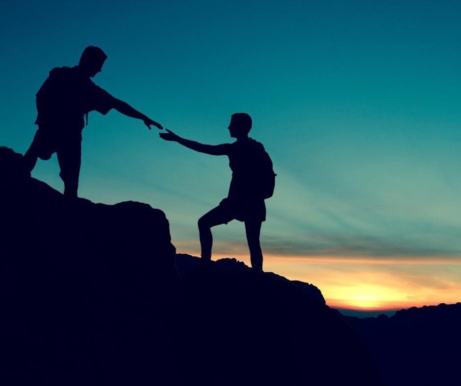 Two silhouetted mountain climbers, one outstretching a hand to thelp the other, in front of a beautiful sunset. 