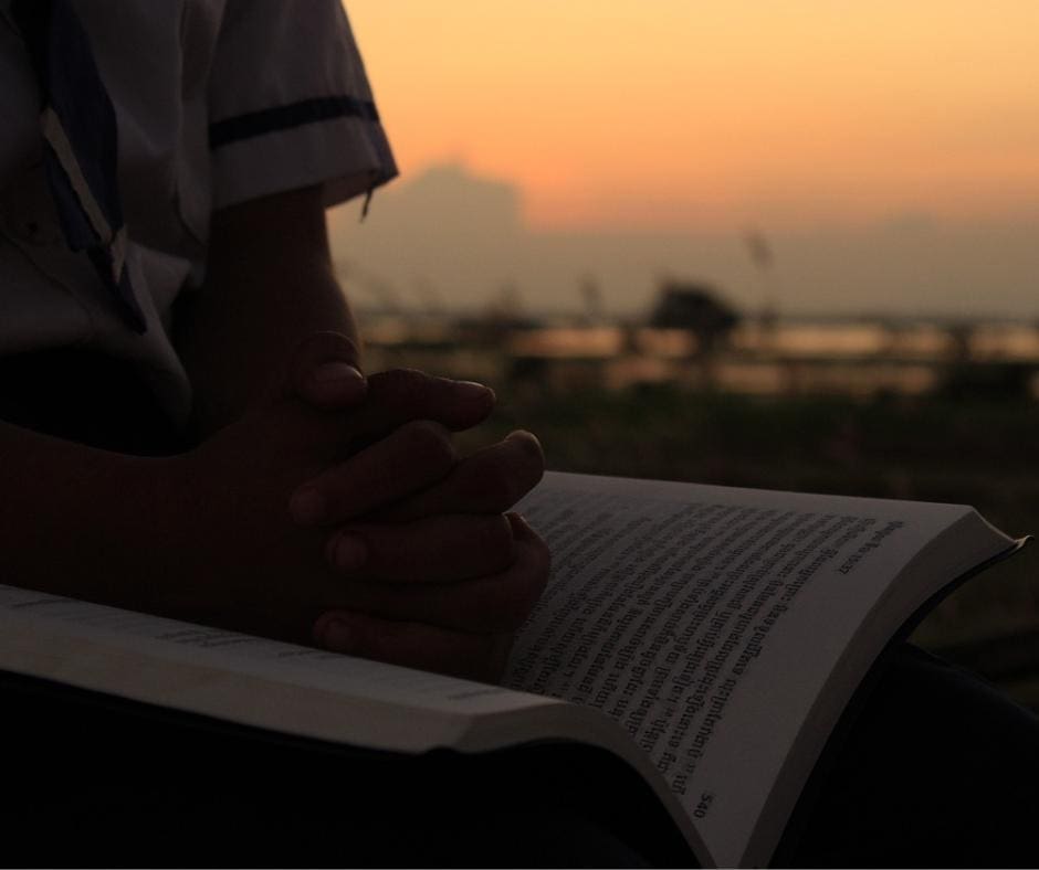 Person with hands folded over a book with Sanskrit writing, sitting in front of a dusty sunset. 