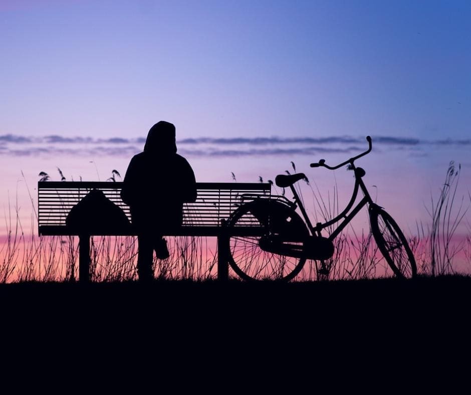 Photo of silhouette of person on park bench with a bicylce parked next to them, watching a beautiful purple and pink sunset in the sky. 