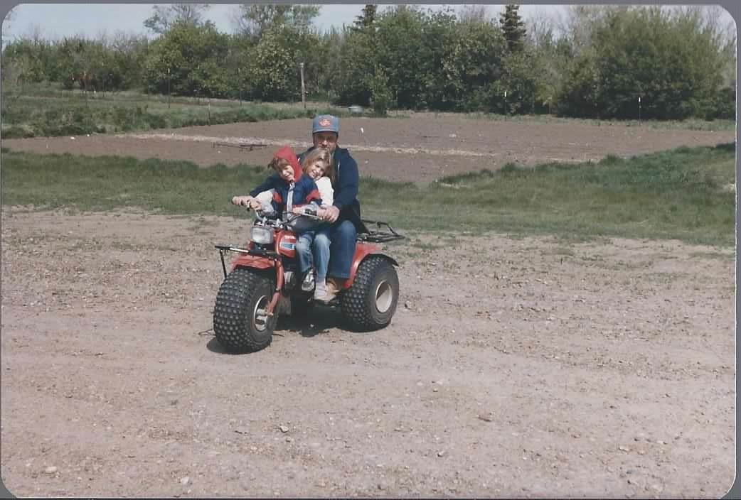 Donna and Michael as children on a three-wheeler with Grandpa at the farm.