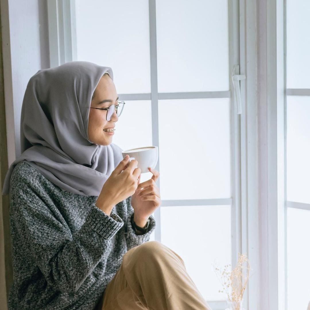 Woman in hijab sitting in window enjoying a cup of tea, practicing mindfulness and self care