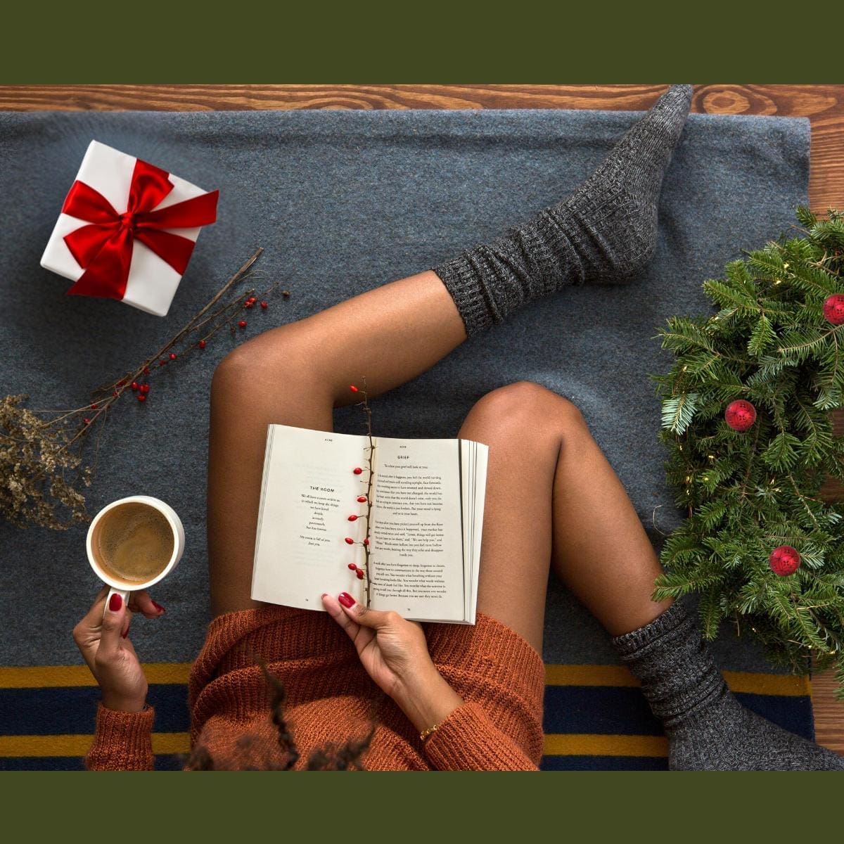 Black female in cozy socks and sweater sitting on blanket with cup of coffee and cozy book amidst holiday gifts and decorations